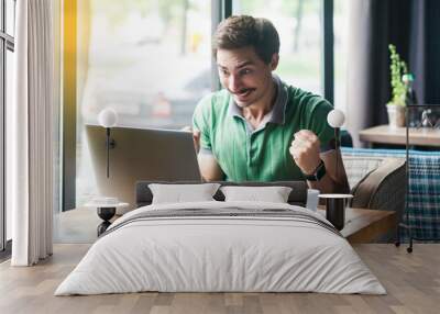 I am winner! Young happy surprised businessman in green t-shirt sitting, looking at laptop screen and rejoicing his success. business and freelancing concept. indoor shot near big window at daytime. Wall mural