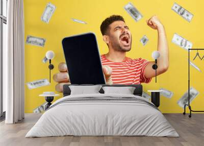 Money rain, winner and rich. Portrait of rejoicing happy bearded man wearing red T-shirt standing with smart phone in hands, showing blank screen. Indoor studio shot isolated on yellow background. Wall mural