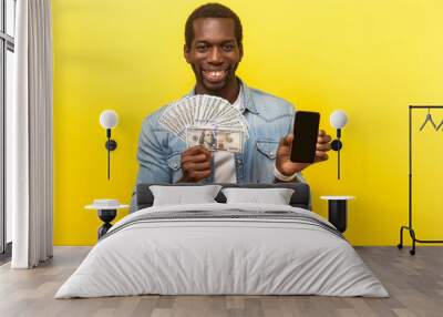 Online banking. Portrait of cheerful young man in denim shirt holding dollars and smartphone, smiling at camera, making money in mobile application. indoor studio shot isolated on yellow background Wall mural