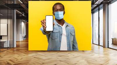 Online service, technology. Portrait of satisfied glad young man with medical mask standing holding out cellphone and smiling broadly at camera. indoor studio shot isolated on yellow background Wall mural