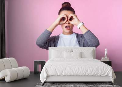 Portrait of amazed brunette teenage girl with bun hairstyle in casual clothes looking through fingers in binoculars gesture, surprised shocked by what she sees. studio shot isolated on pink background Wall mural