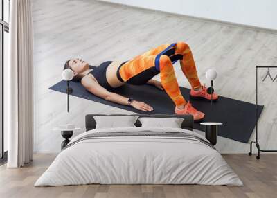 Portrait of athletic woman in bridge pose using elastic band for legs and butt workout, body care, wearing black tank top and orange leggings. Full length indoor shot against white wall. Wall mural