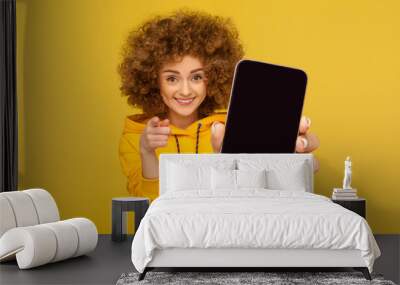 Portrait of attractive smiling woman with Afro hairstyle wearing hoodie showing big mobile display for copy space and pointing to camera, choosing you. Indoor studio shot isolated on yellow background Wall mural