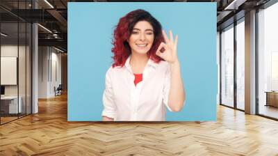 Portrait of cheerful adult woman with fancy red hair showing okay sign, assures you everything is fine, looks gladly, wearing white shirt. Indoor studio shot isolated on blue background. Wall mural