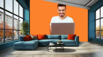 Portrait of cheerful young brunette man with beard in white t-shirt typing email on laptop and smiling, reading positive message or surfing the web. indoor studio shot isolated on orange background Wall mural