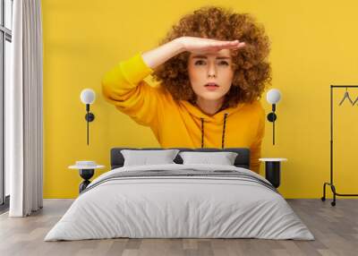 Portrait of curious curly-haired woman in urban style hoodie holding hand above eyes and peering into distance, looking far away, expecting and searching someone on horizon. studio shot isolated Wall mural