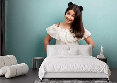 Portrait of happy beautiful brunette young girl with black long hair in white dress standing with hands on waist and looking at camera with toothy smile. indoor studio shot isolated on blue background Wall mural