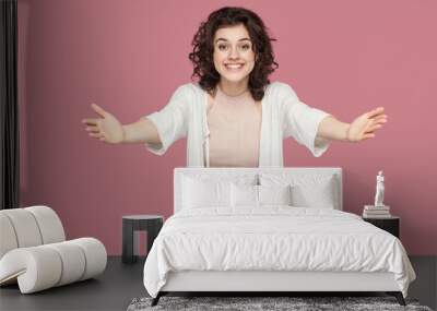 Portrait of happy beautiful brunette young woman with curly hairs in casual style standing with raised arms, want to hug, toothy smile and looking at camera. studio shot isolated on pink background. Wall mural