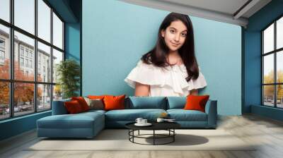 Portrait of happy calm beautiful brunette young girl with black long straight hair in white dress standing and looking at camera with smile. indoor studio shot isolated on blue background. Wall mural