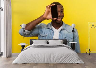 Portrait of happy cheerful man in denim casual shirt covering half face with cellphone and looking at camera with toothy smile, positive glad face. indoor studio shot isolated on yellow background Wall mural