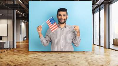 Portrait of happy positive man with beard standing and pointing at american flag, celebrating national holiday, wearing striped shirt. Indoor studio shot isolated on blue background. Wall mural