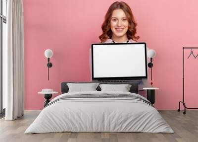 Portrait of happy smiling curly haired teenage girl in hoodie showing empty laptop screen on camera, place for advertisement. Indoor studio shot isolated on pink background Wall mural