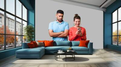 Portrait of jealous boyfriend standing with crossed arms, looking suspicious at woman cheating using phone and smiling happily, unfaithful relations. isolated on gray background, indoor studio shot Wall mural