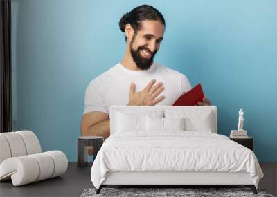 Portrait of man with beard wearing T-shirt reading letter from red envelope, gets greeting card, having positive romantic expression, being pleased, Indoor studio shot isolated on blue background. Wall mural