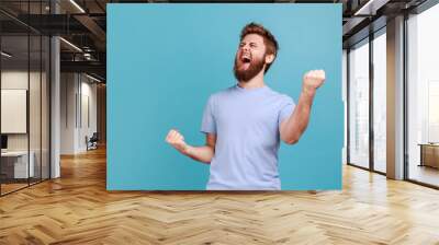 Portrait of overjoyed bearded man standing with excited expression, raising fists, screaming, shouting yeah, celebrating his victory, success. Indoor studio shot isolated on blue background. Wall mural