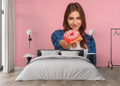 Portrait of positive girl in checkered shirt holding out tasty donut and smiling to camera, showing sweetened confectionery high in glucose and calories. indoor studio shot isolated on pink background Wall mural