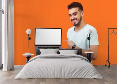 Portrait of positive joyful brunette man with beard in white t-shirt standing holding laptop with blank screen and smiling at camera, internet advertising. studio shot isolated on orange background Wall mural