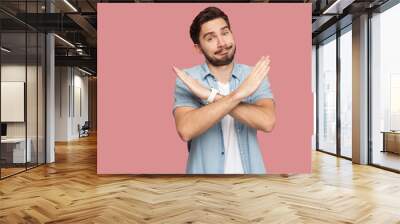 Portrait of serious handsome bearded young man in blue casual style shirt standing with X sign hands and looking at camera. indoor studio shot, isolated on pink background. Wall mural