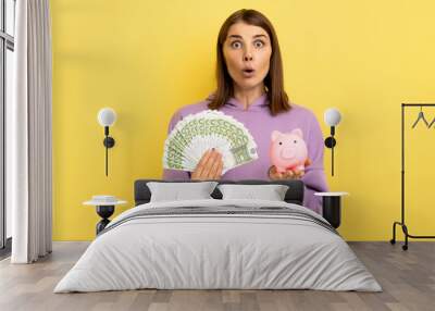 Portrait of shocked amazed woman with dark hair holding big sum of money and piggy bank, profitable investment, wearing purple hoodie. Indoor studio shot isolated on yellow background. Wall mural