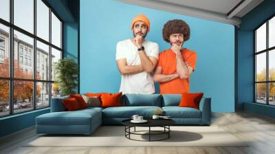 Portrait of two young adult hipster men holding chin with hands, need to think, planning strategy, thinking over ideas for startup. Indoor studio shot isolated on blue background. Wall mural