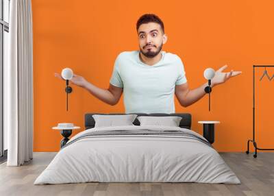 Portrait of uncertain puzzled young bearded man wearing T-shirt shrugging shoulders spreading arms doesn't know answer. Indoor studio shot isolated on orange background. Wall mural