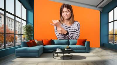 Portrait of upset woman with brown hair in long sleeve shirt standing with grimace of pain, massaging sore wrist, suffering hand injury or sprain. indoor studio shot isolated on orange background Wall mural