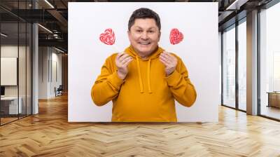 Positive satisfied man with dark hair holding two heart shape lollipops, having fun, positive expressions, wearing urban style hoodie. Indoor studio shot isolated on white background. Wall mural