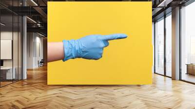 Profile side view closeup of human hand in blue surgical gloves showing or pointing ot something with finger. indoor, studio shot, isolated on yellow background. Wall mural