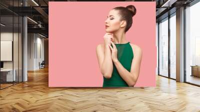 Profile side view portrait of calm serious beautiful young woman with bun hairstyle and makeup in green dress standing with closed eyes and touching her chin. studio shot, isolated on pink background. Wall mural