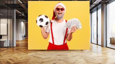 Satisfied elderly man in santa claus hat and stylish sunglasses holding soccer ball and dollar banknotes, sports betting, big win. Indoor studio shot isolated on yellow background Wall mural