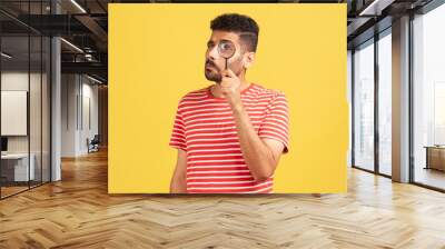 Serious bearded man in red striped t-shirt standing, holding magnifying glass and looking at camera with big zoom eye, verifying authenticity Wall mural