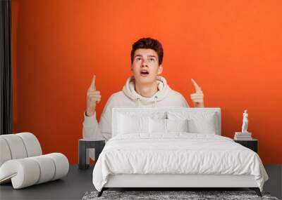 Shocked teenager in white sweater with hood pointing fingers up and looking with widely opened eyes and mouth, amazed with your advertising banner. Indoor studio shot isolated on orange background Wall mural