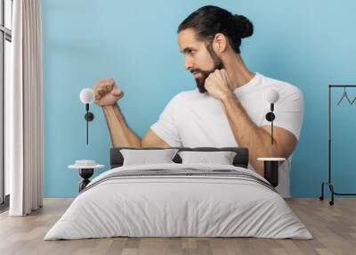 Side view of aggressive man with beard wearing white T-shirt holding clenched fists up ready to boxing, martial art trainer, self defense. Indoor studio shot isolated on blue background. Wall mural