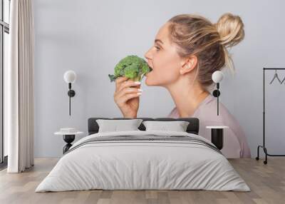 Side view of attractive young woman with hair bun biting green broccoli, eating fresh vegetable, enjoying healthy food, vegetarian diet, nutrition. indoor studio shot isolated on grey background Wall mural