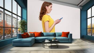 Side view of surprised teenager girl in yellow casual T-shirt, expressing shock while using cell phone, searching web, astonishing news. Indoor studio shot isolated on gray background. Wall mural