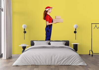 Side view portrait of courier woman standing with two parcels in hands, looking ahead, delivery service, wearing overalls and red cap. Indoor studio shot isolated on yellow background. Wall mural