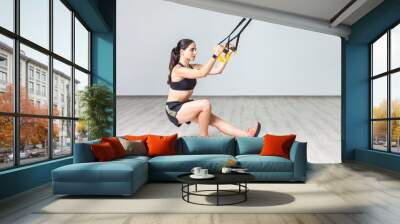 Side view portrait of muscular beautiful woman wearing sportswear squatting with fitness straps in the gym, using trx for her body workout. Indoor shot against white wall background Wall mural