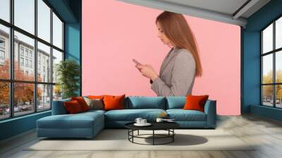 Side view portrait of serious woman with brown hair using mobile phone, typing messages, surfing internet, wearing business suit. Indoor studio shot isolated on pink background. Wall mural