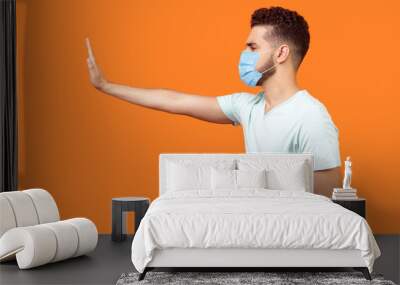 Stop, don't come any closer. Side view of serious bossy man with surgical medical mask standing holding out hand to left, prohibition gesture, saying no. studio shot isolated on orange background Wall mural