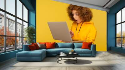 Surprised emotional bright woman with curly hair in urban style hoody looking at laptop screen with shocked expression, typing and browsing internet. indoor studio shot isolated on yellow background Wall mural