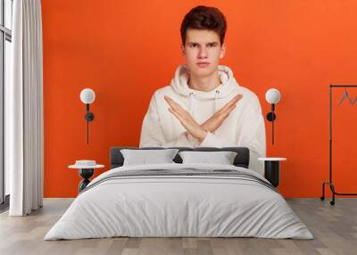 There is no way! Serious concentrated young man in stylish hoodie showing no gesture crossing arms, say no to bad habits, addiction. Indoor studio shot isolated on orange background Wall mural