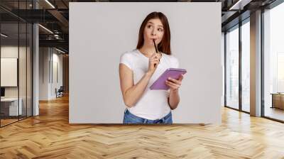 Thoughtful woman standing with paper notebook and pen, looking away with pensive expression, making to do list, wearing white T-shirt. Indoor studio shot isolated on gray background. Wall mural