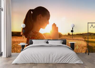 Young woman meditating, praying with her eyes closed, practicing self awareness with hands in prayer position against a beautiful sunset. 
 Wall mural