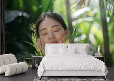 A beautiful asian woman with her eyes closed stands in front of green plants and palm trees, holding both hands to her face for skin care Wall mural