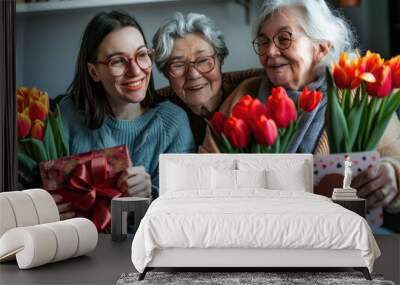 A happy grandmother, mother and daughter sitting on the sofa at home holding flowers tulips in one hand, an open red gift box with chocolates inside it, they have cards to write love messages for Moth Wall mural