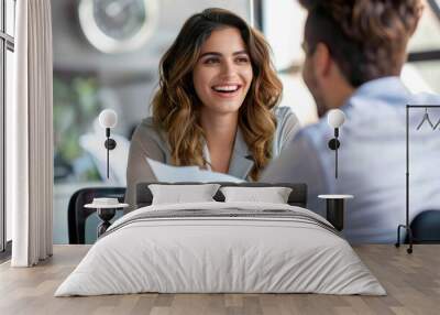 a happy young woman sitting at an office desk, holding papers and smiling as she talks to her male hopeful employee during the interview process Wall mural