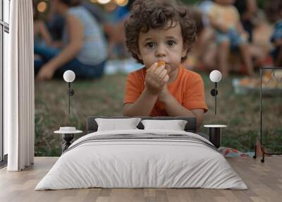 A little boy sits on the lawn in an orange T-shirt and gray pants, eating chips from his hand with his fingers. People are behind him at a summer picnic at sunset in the park Wall mural