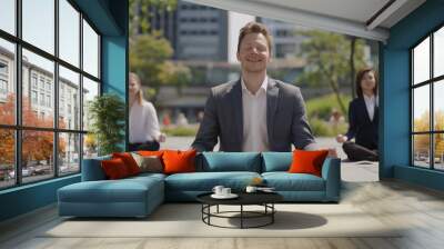 A smiling businessman in business attire meditating outdoors with other happy office workers, sitting on the ground doing yoga poses Wall mural