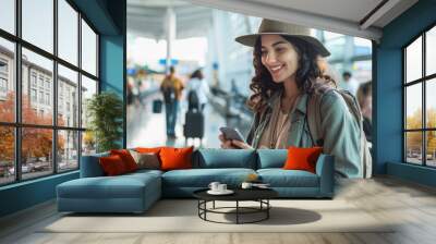 A young woman in casual attire, wearing an elegant hat and carrying travel backpack is standing at the airport waiting for her flight Wall mural