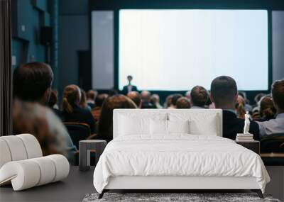 an audience from the front and side views, with people sitting in chairs watching someone on stage presenting to them. A white screen for presentations is on a big wall behind the conference room Wall mural